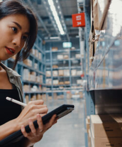 Eine Frau schreibt auf einem Tablet in einem Lager. Sie steht zwischen Regalen mit Kartons und konzentriert sich auf ihre Arbeit.