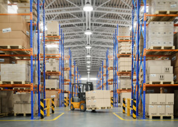 Ein Gabelstapler fährt durch ein gut organisiertes Lager und transportiert Kartons auf Regalen.