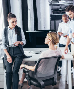 Ein Büro-Team führt ein Gespräch. Eine Person steht und hält ein Tablet, während eine andere auf einem Bürostuhl sitzt. Zwei weitere Teammitglieder sind im Hintergrund aktiv.