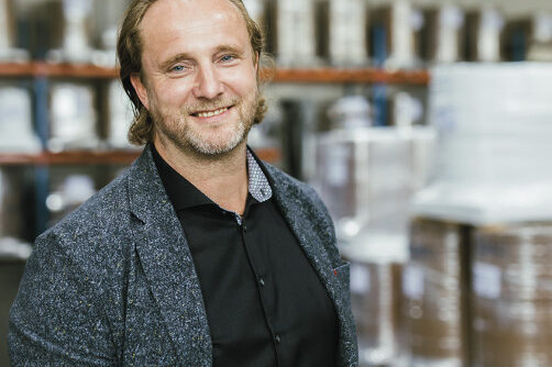 Porträt eines Mannes in einem Lagerumfeld, er lächelt und trägt ein dunkelblaues Hemd und einen grauen Blazer. Im Hintergrund sind Paletten mit Waren sichtbar.