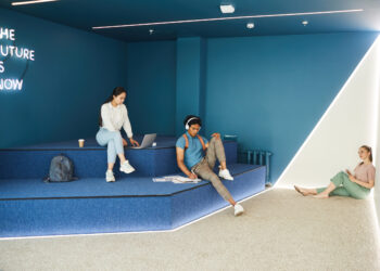 Moderner Studienraum mit leuchtend blauen Wänden, ein Student arbeitet am Laptop, ein zweiter Student entspannt beim Lesen.